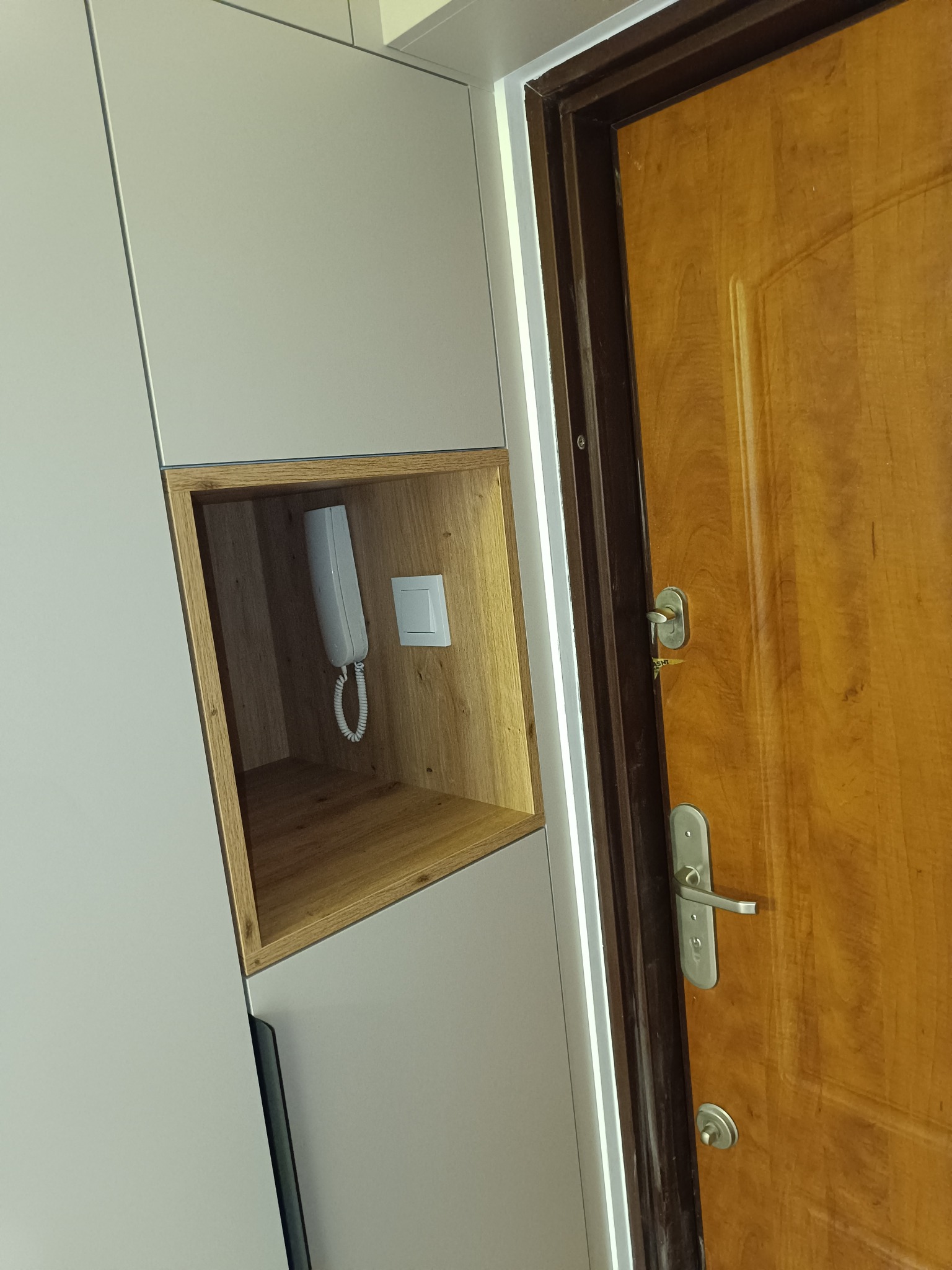 Biała szafa wnękowa z otwartą półką z drewnianym wykończeniem, w której znajduje się biały telefon stacjonarny i włącznik światła. Obok widoczna drewniana drzwi.