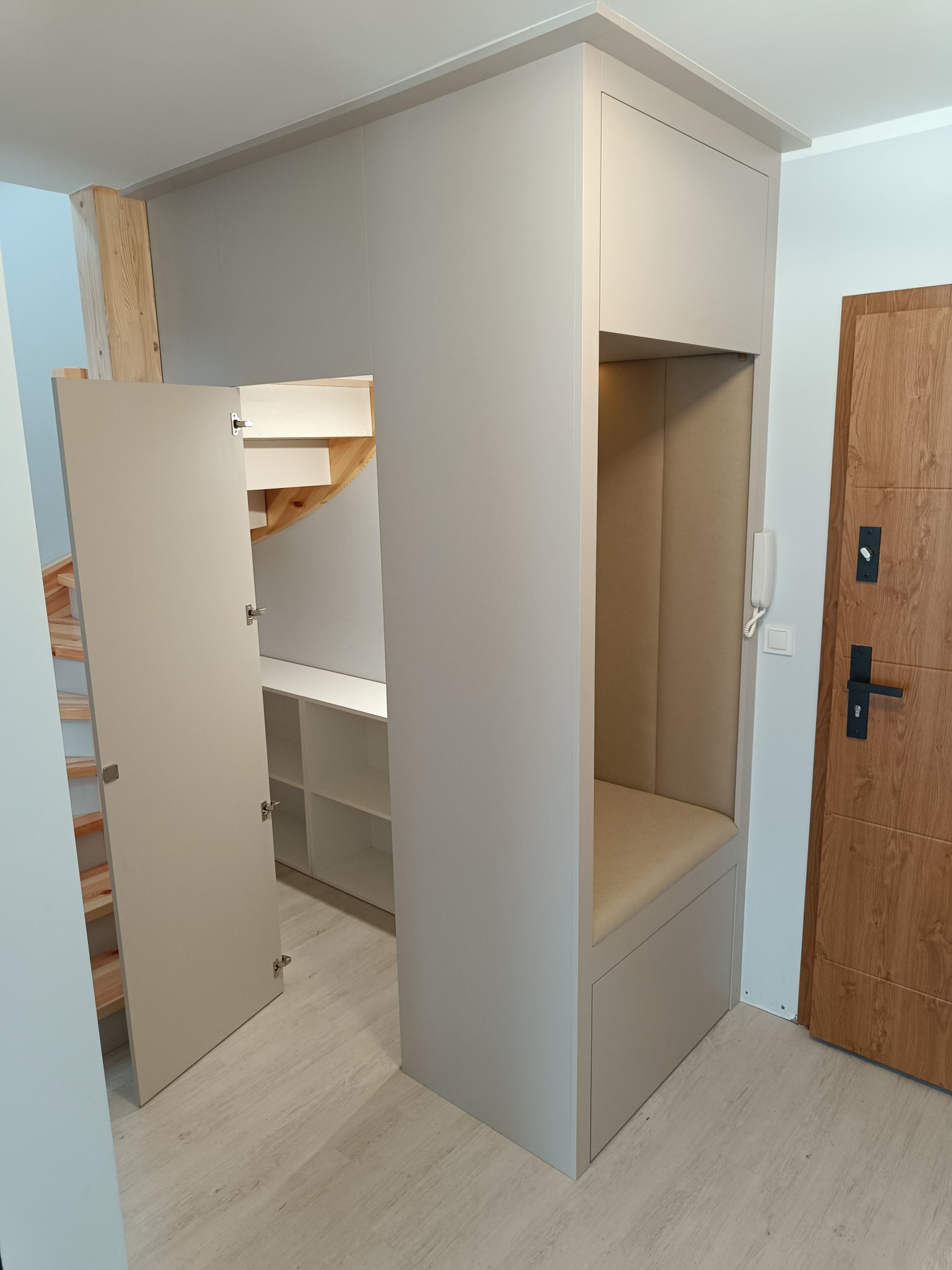 Szara zabudowa wnęki pod schodami z otwartą szafką i siedziskiem. Widoczne półki wewnątrz. Drzwi wejściowe po prawej stronie.