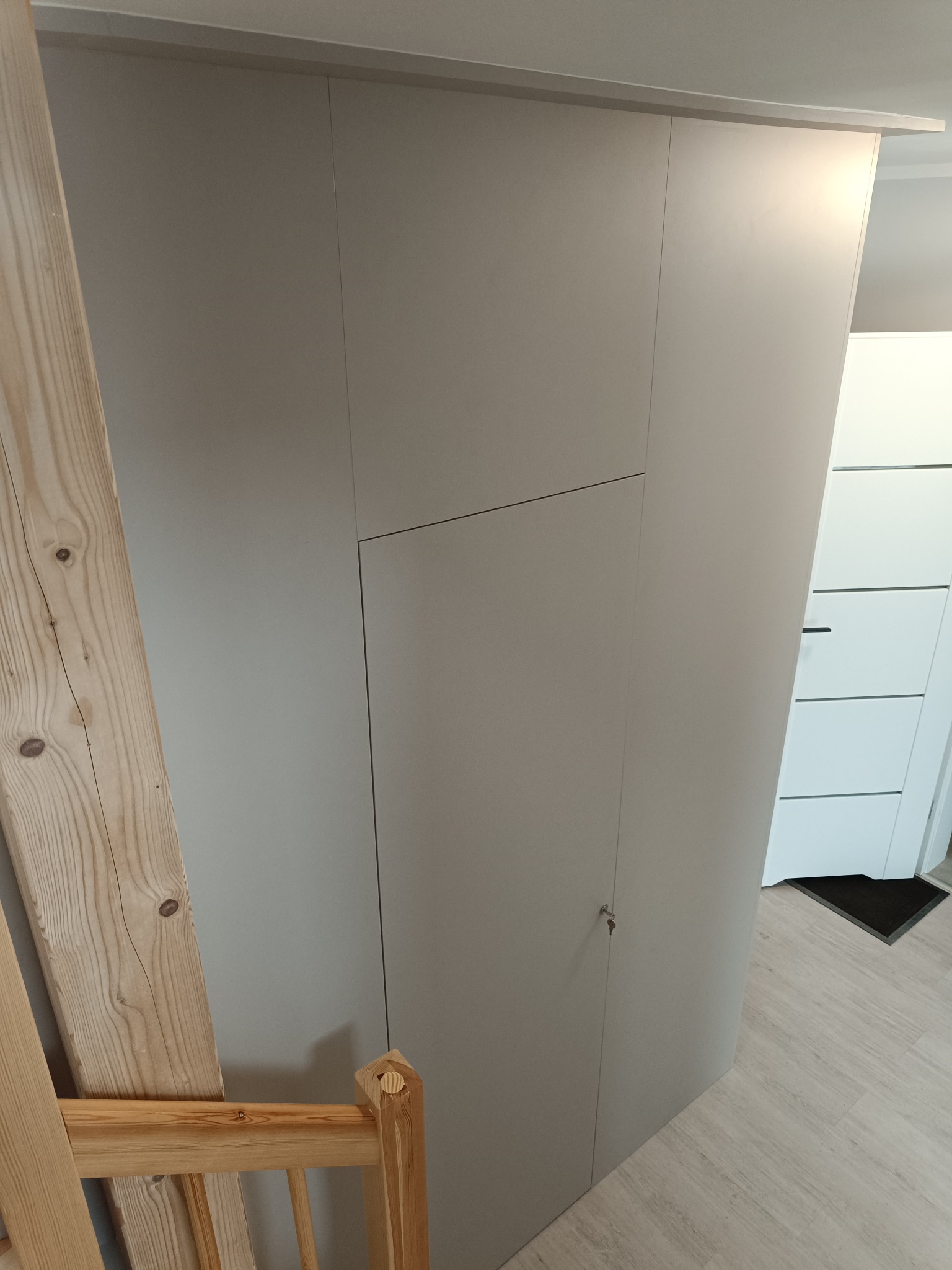 Jasnoszara szafa wnękowa z małym zamkiem, widoczna z perspektywy podłogi, z elementami drewnianymi w kadrze. Minimalistyczny design.