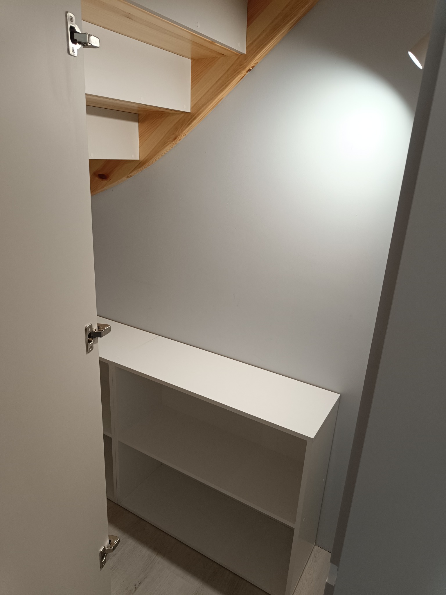 Biała szafka z półkami pod schodami, widoczna z otwartymi drzwiami szafy. Drewniane schody i szara ściana w tle. Oświetlenie punktowe podkreśla strukturę ściany.