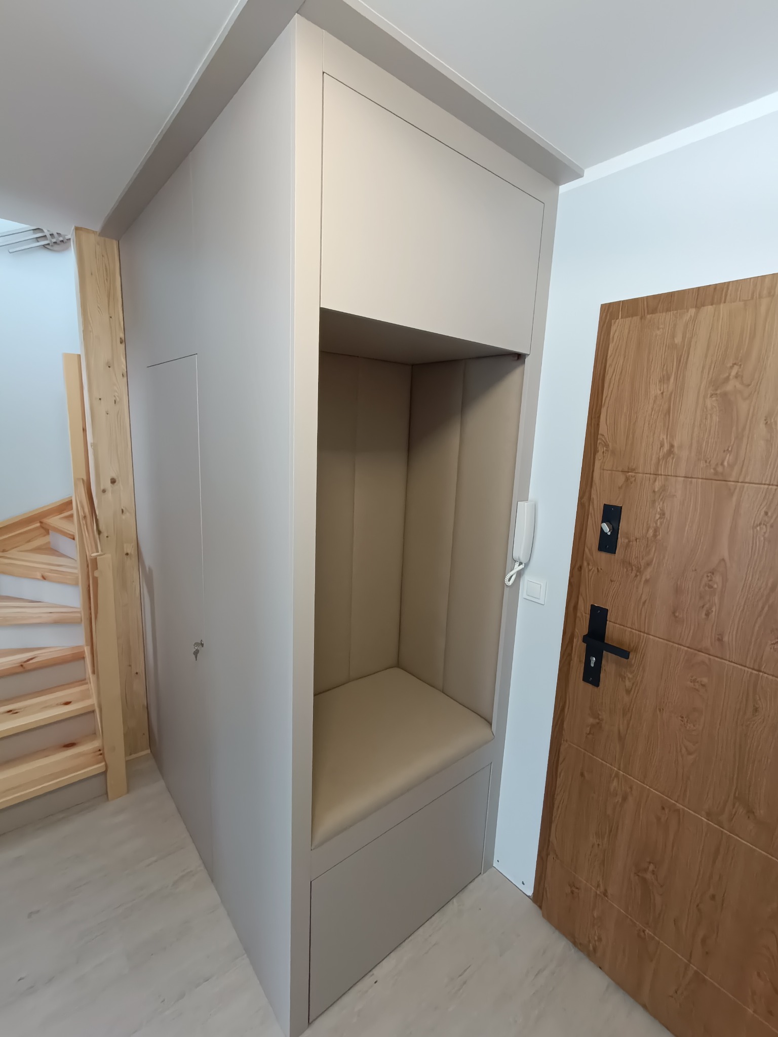 Szara zabudowa w przedpokoju z siedziskiem i szafką, obok drewniane schody i drzwi wejściowe. Minimalistyczny design, jasne wnętrze.