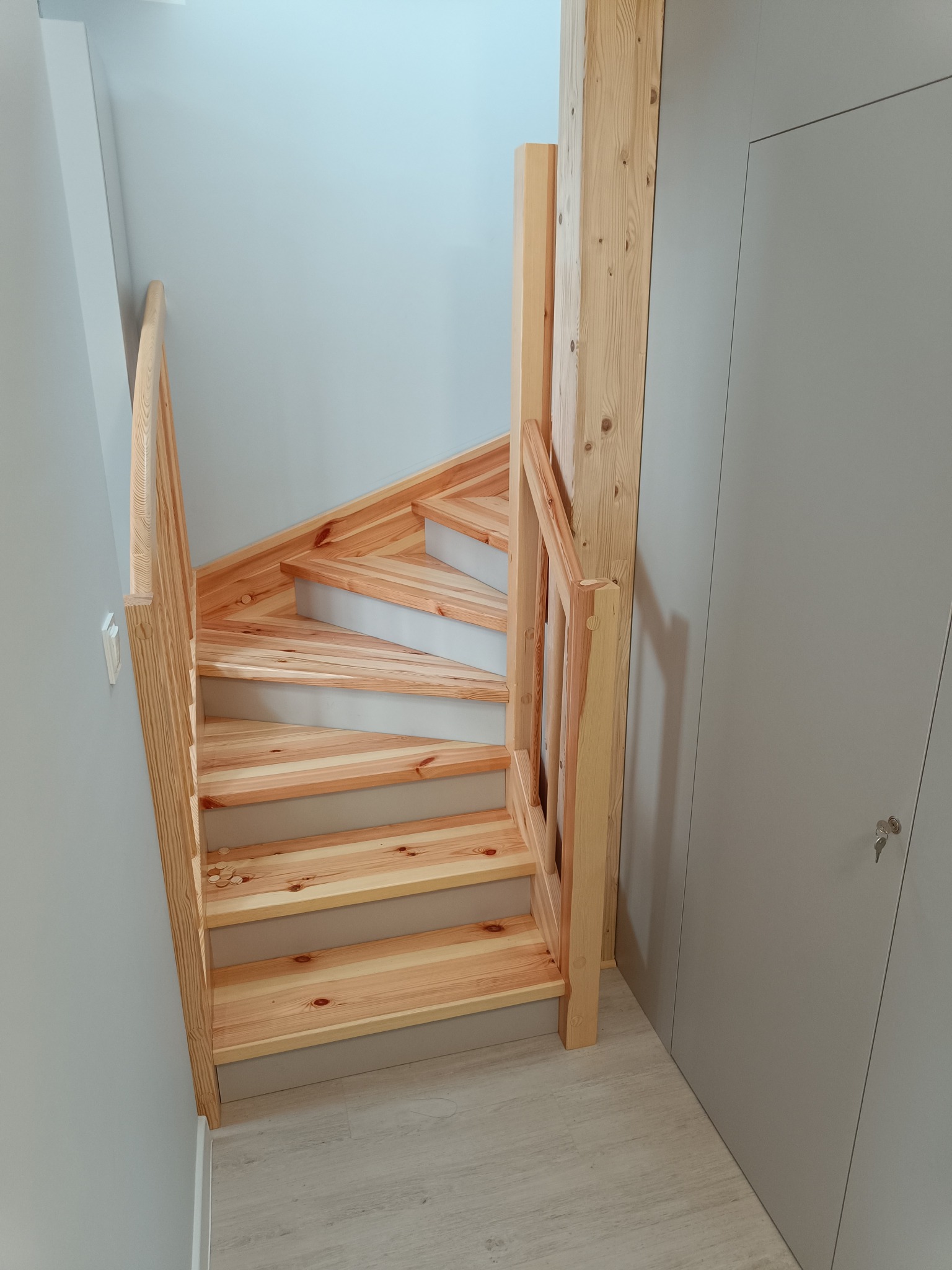 Drewniane schody z jasnego drewna sosnowego, prowadzące w górę, z szarą ścianą i drzwiami szafy w tle. Widok z dołu, podkreślający naturalny rysunek drewna.