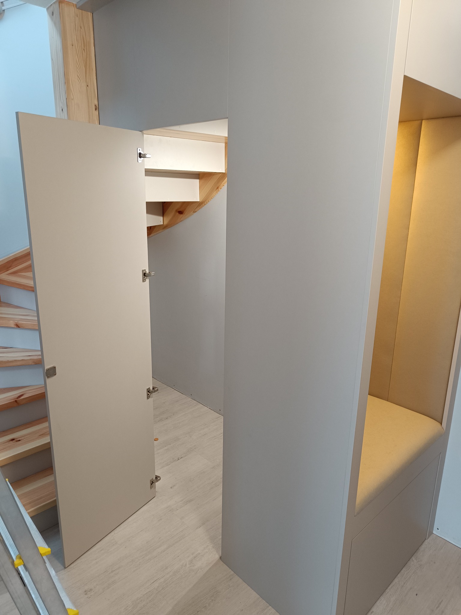 Biała szafa wnękowa z siedziskiem i schodami z jasnego drewna. Otwarta szafka ukazuje wnętrze z półkami. Widoczny fragment drabiny.