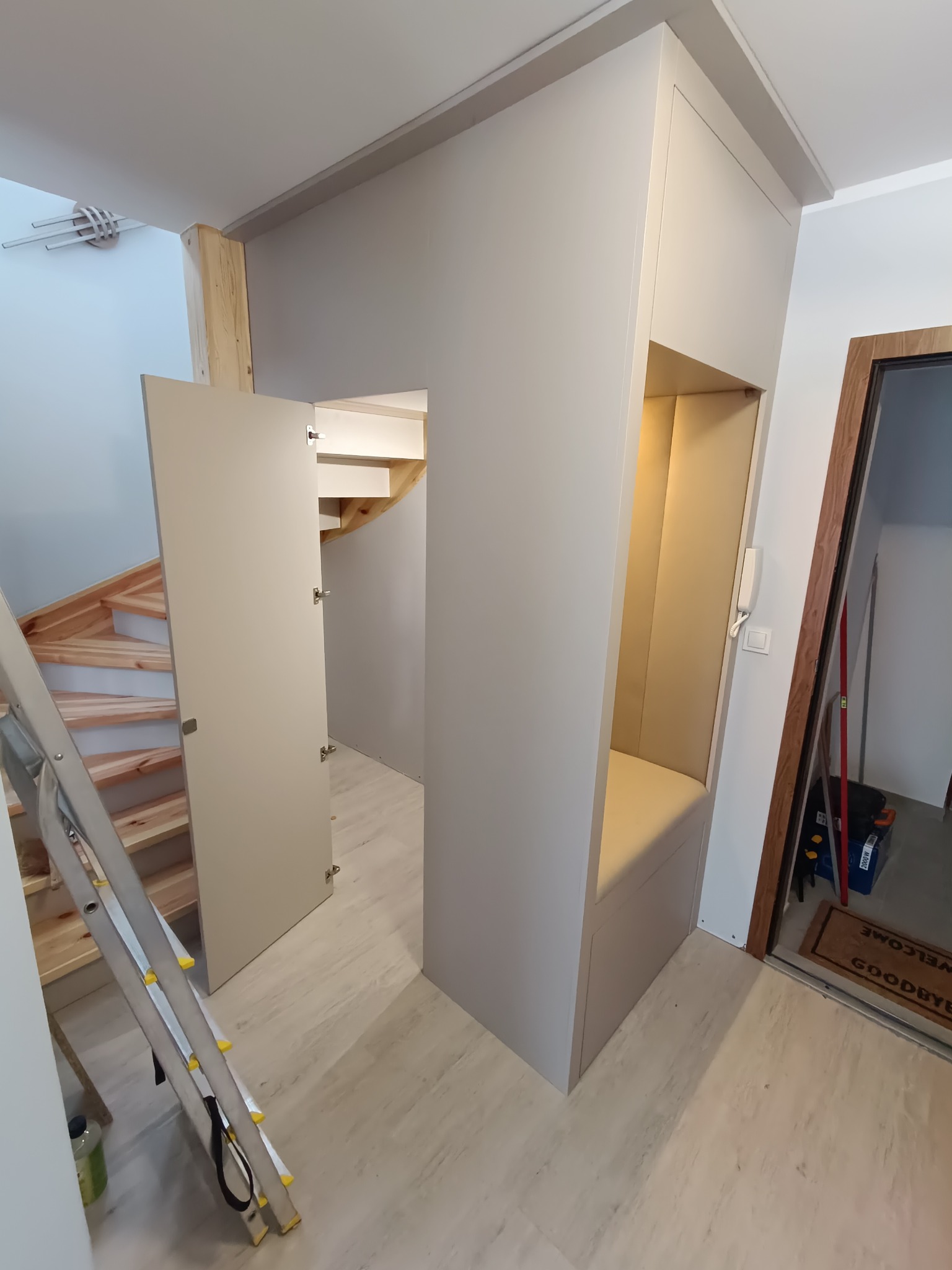 Szara szafa wnękowa z siedziskiem i otwartą przestrzenią, wkomponowana w przestrzeń pod schodami. Widoczne schody drewniane i drabina aluminiowa.