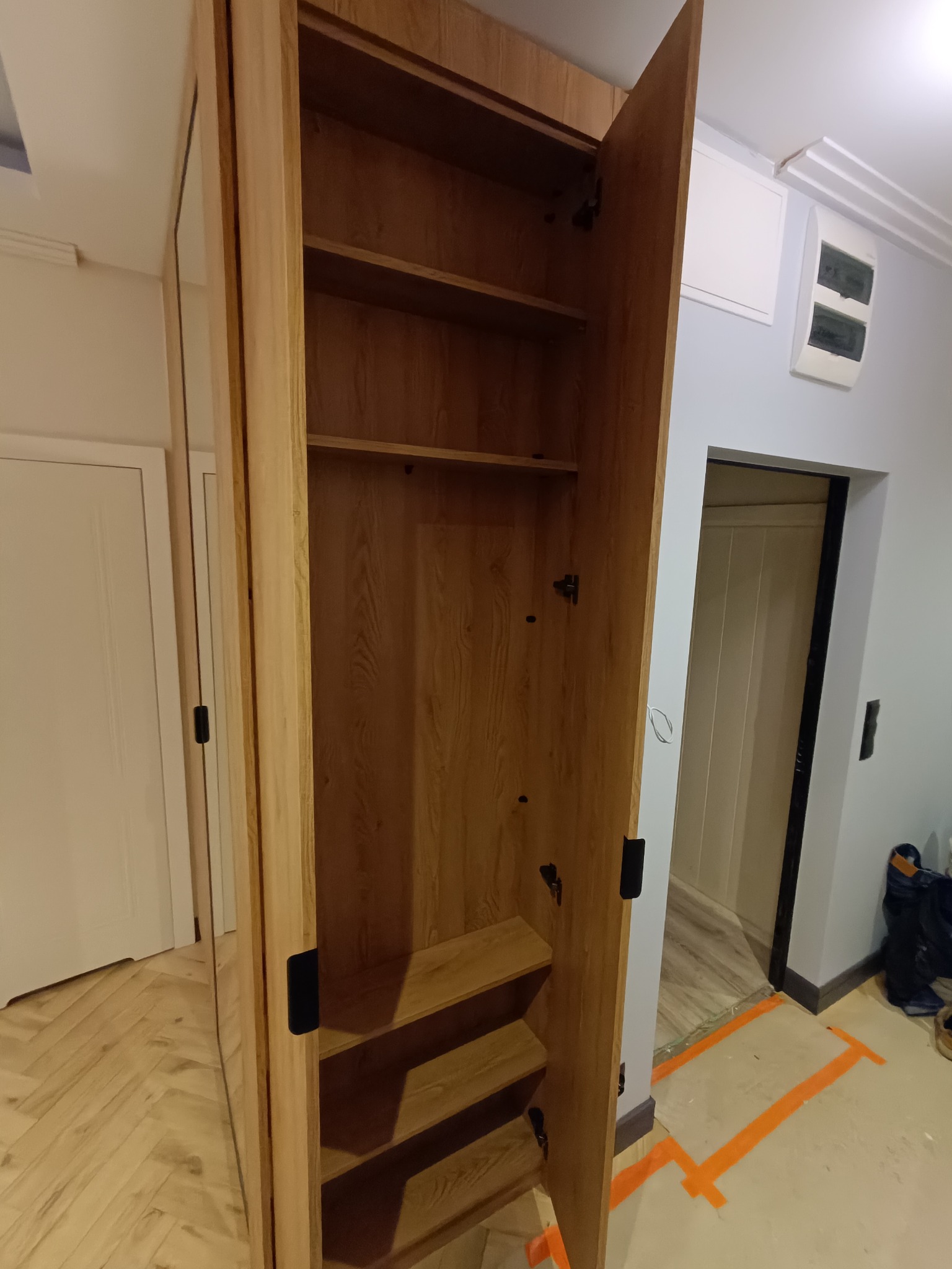 Otwarta, drewniana szafa z półkami i lustrzanym frontem, widoczna w jasnym wnętrzu z parkietem ułożonym w jodełkę. Drzwi szafy otwarte, ukazują wnętrze z półkami.