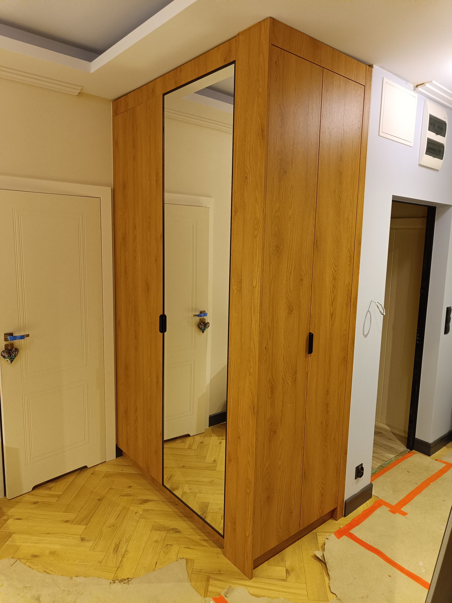 Drewniana szafa wnękowa z lustrem, czarne uchwyty, parkiet w jodełkę. Widoczne zabezpieczenia podłogi. Wnętrze szafy odbija pokój.