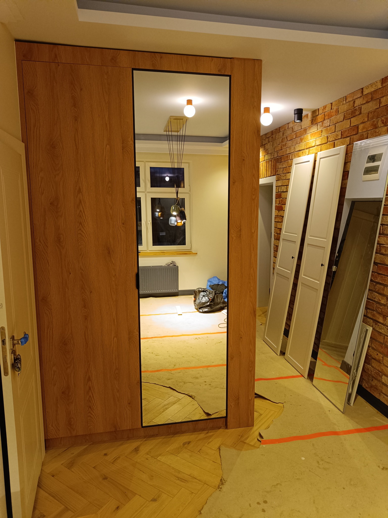 Montaż szafy wnękowej z lustrem w trakcie wykańczania wnętrza. Widoczne zabezpieczenia podłogi i ściany z cegły. Częściowo zamontowane drzwi do szafy oparte o ścianę.