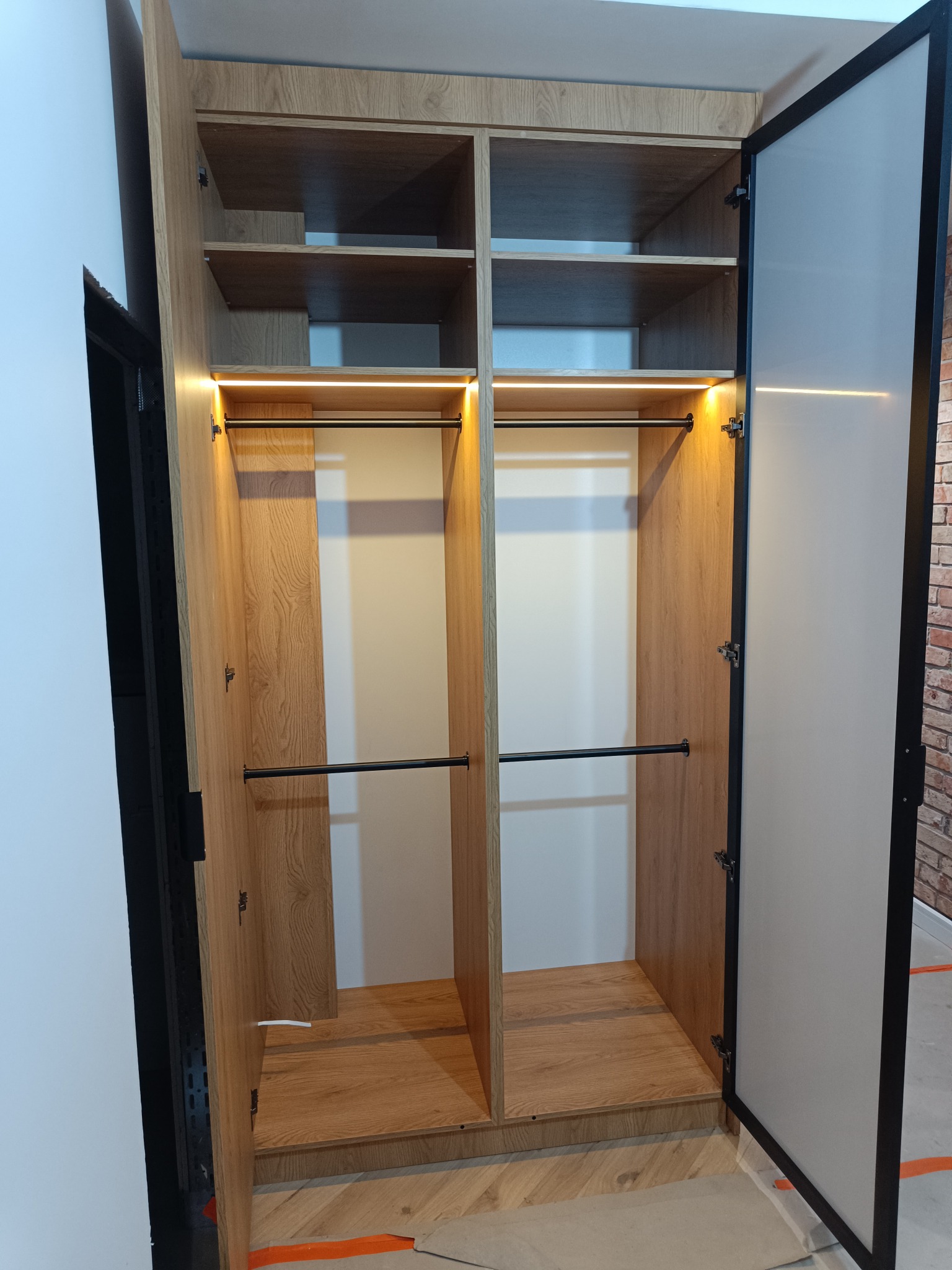 Otwarta szafa wnękowa z oświetleniem LED, drewnopodobne wykończenie, czarne relingi, widoczne półki i elementy montażowe. Drzwi z czarną ramą.