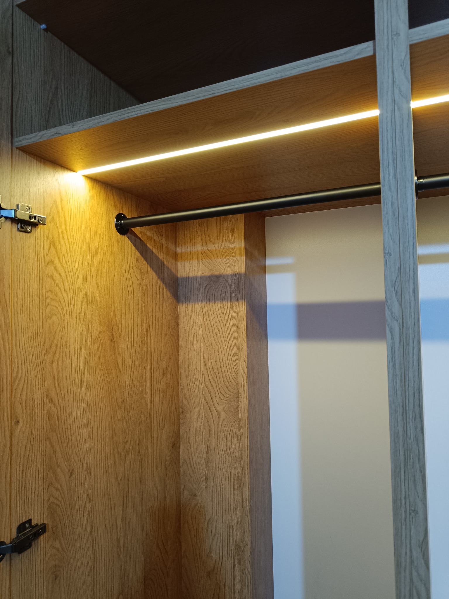 Wnętrze szafy wnękowej z oświetleniem LED, widoczny drążek na ubrania i półki z drewnopodobnym wykończeniem. Zawiasy szafki po lewej stronie kadru.