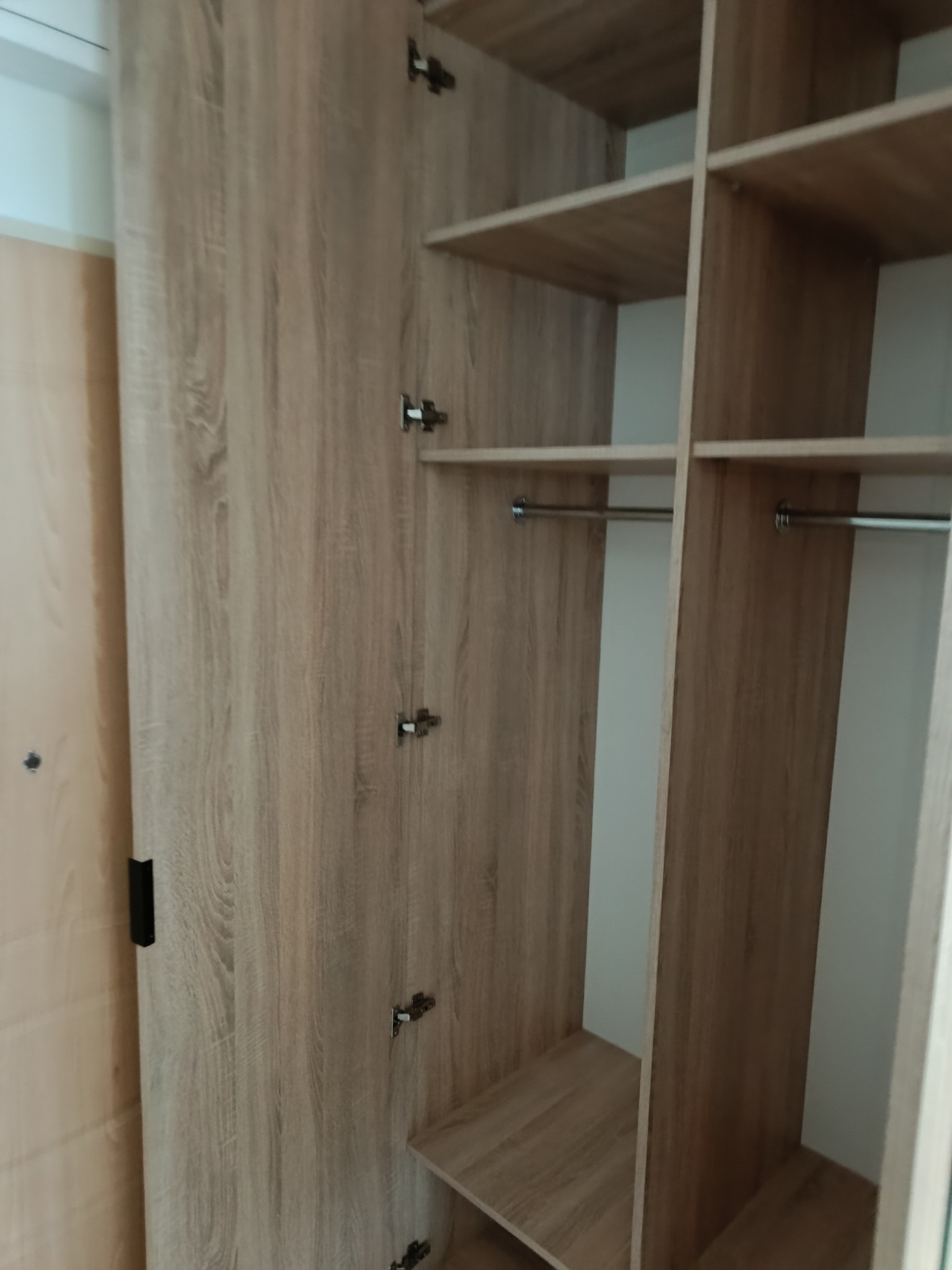 Otwarta szafa wnękowa z półkami i drążkami, wykończona w odcieniach drewna, widoczne zawiasy i elementy montażowe, wnętrze szafy oświetlone.