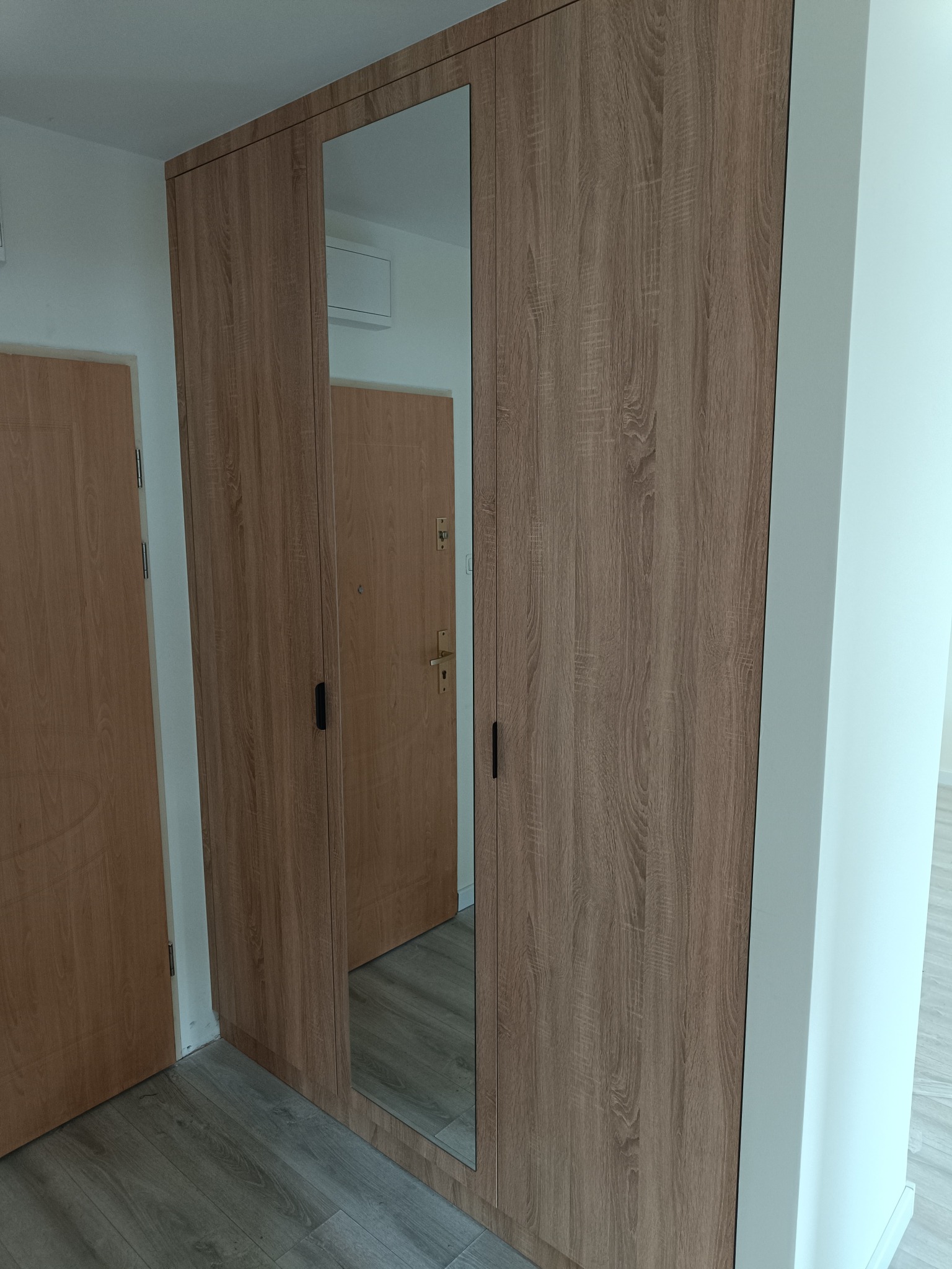 Trzydrzwiowa szafa z lustrem w środkowej części, wykończona okleiną imitującą drewno. Widoczne dwie drewniane drzwi po bokach i fragment podłogi.