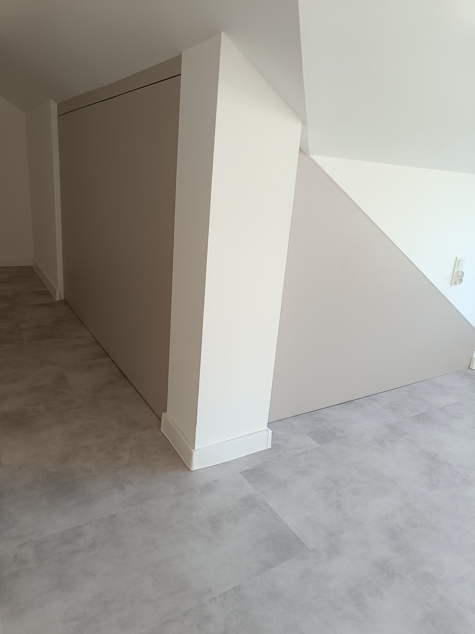 Beżowa szafa wnękowa pod skosem schodów, dopasowana do wnętrza w Dzierżążnie. Minimalistyczny design, szara podłoga, białe wykończenia.