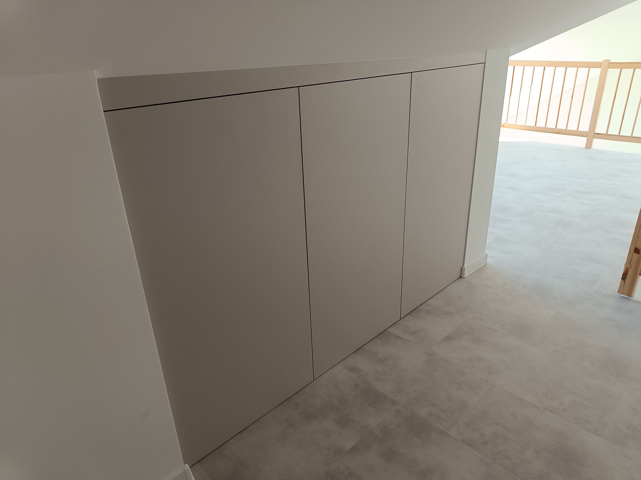 Jasna szafa wnękowa z trzema frontami, wkomponowana w skos poddasza. Minimalistyczny design, szare panele podłogowe, fragment balustrady w tle.