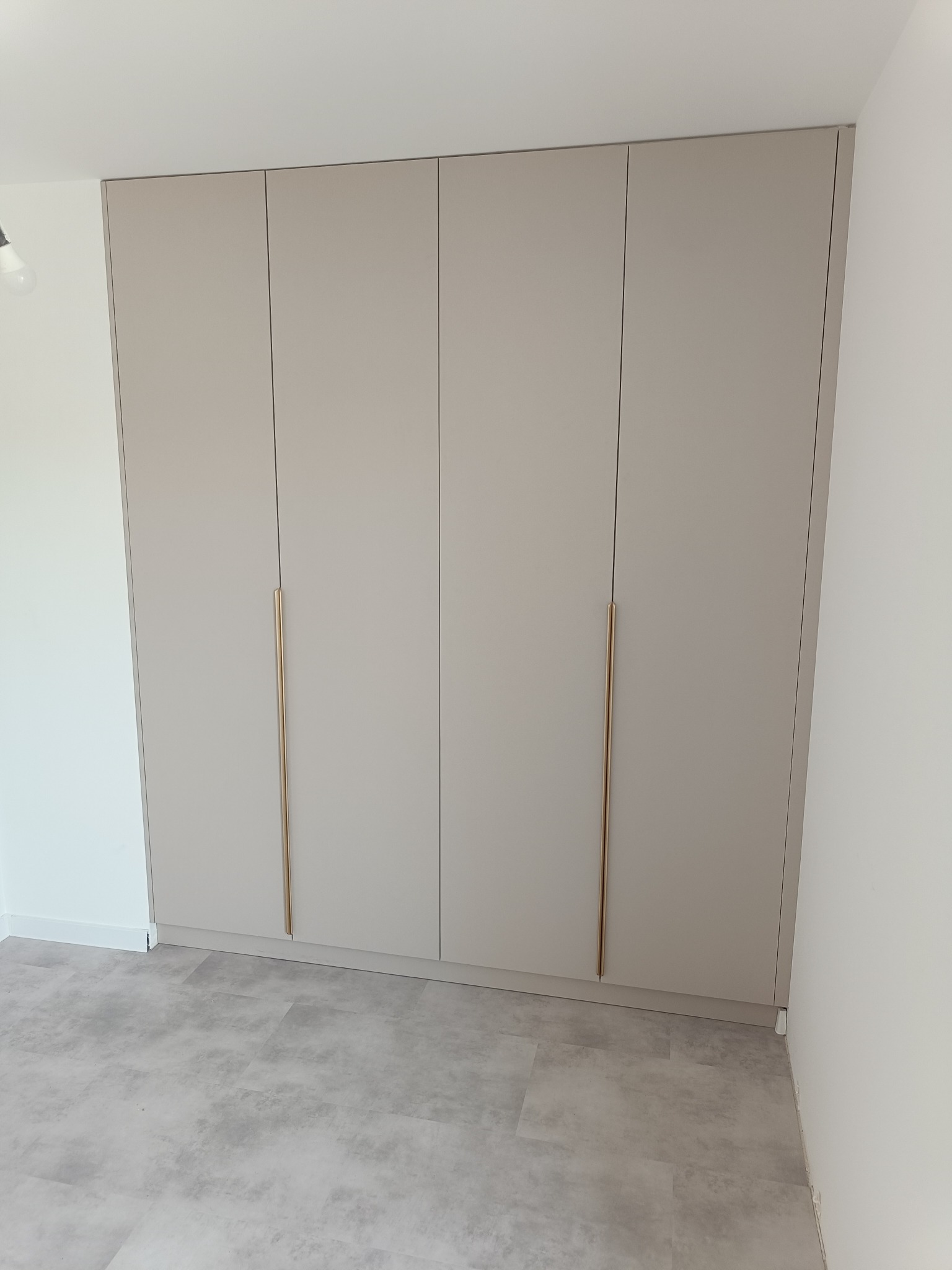 Beżowa szafa wnękowa z czterema drzwiami i złotymi uchwytami, dopasowana do wnętrza z szarą podłogą. Minimalistyczny design, nowoczesne wykończenie.