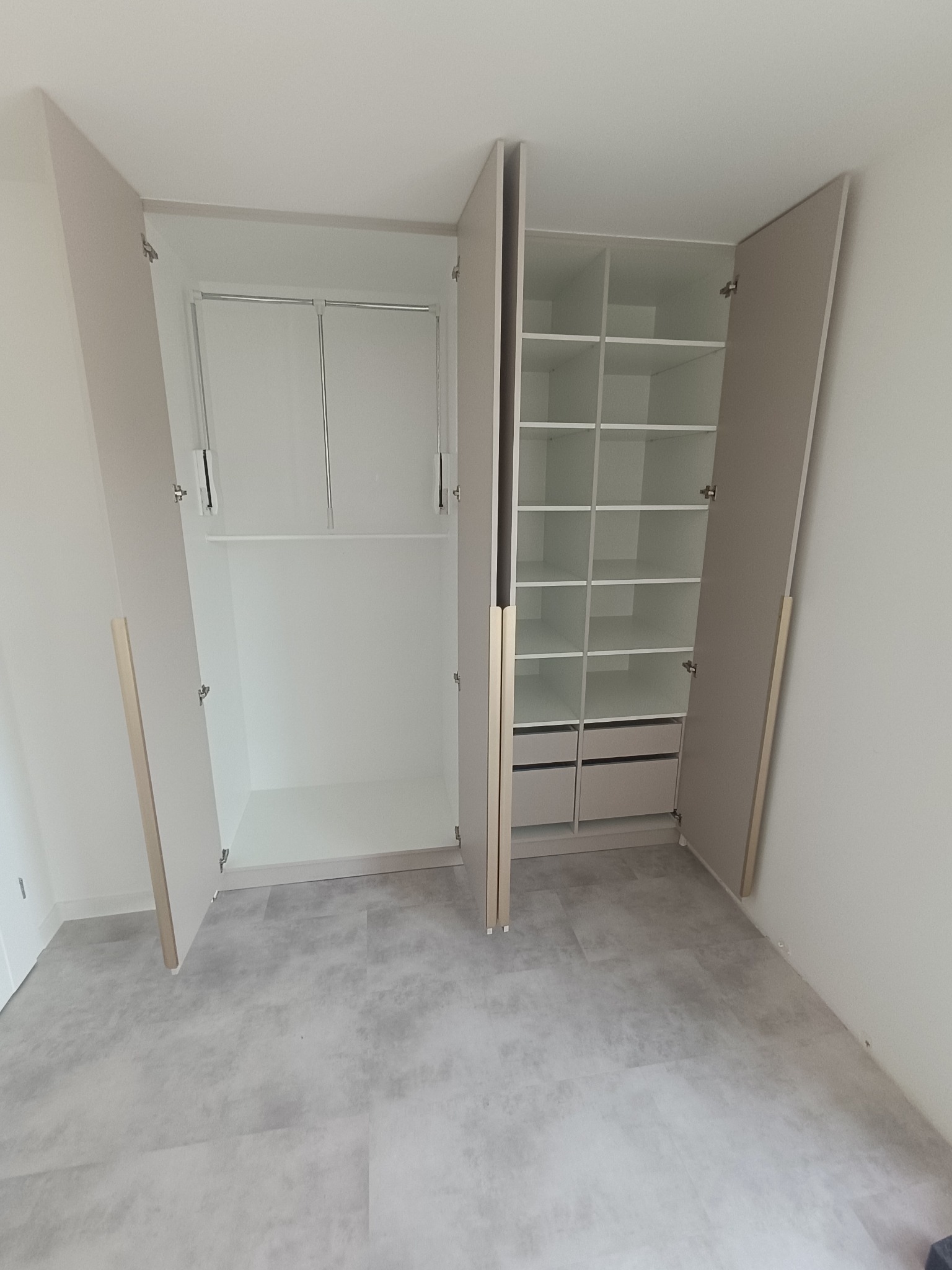 Jasna szafa wnękowa z otwartymi drzwiami, ukazująca półki i drążek na ubrania. Minimalistyczny design, neutralna kolorystyka, szare panele podłogowe.