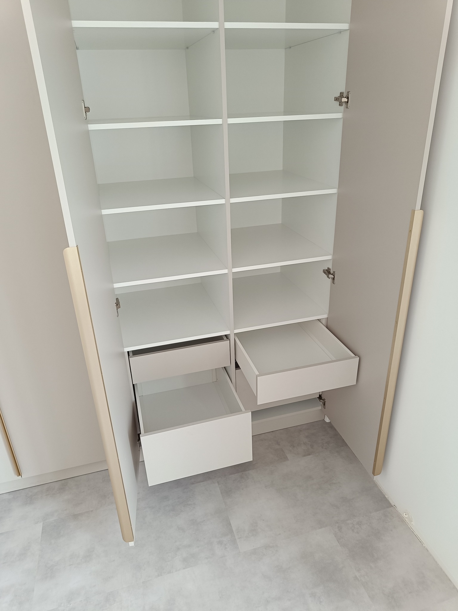 Wnętrze szafy wnękowej z otwartymi drzwiami, widoczne półki i dwie wysunięte szuflady. Jasna kolorystyka, minimalistyczny design. Ujęcie z góry, widoczna podłoga.