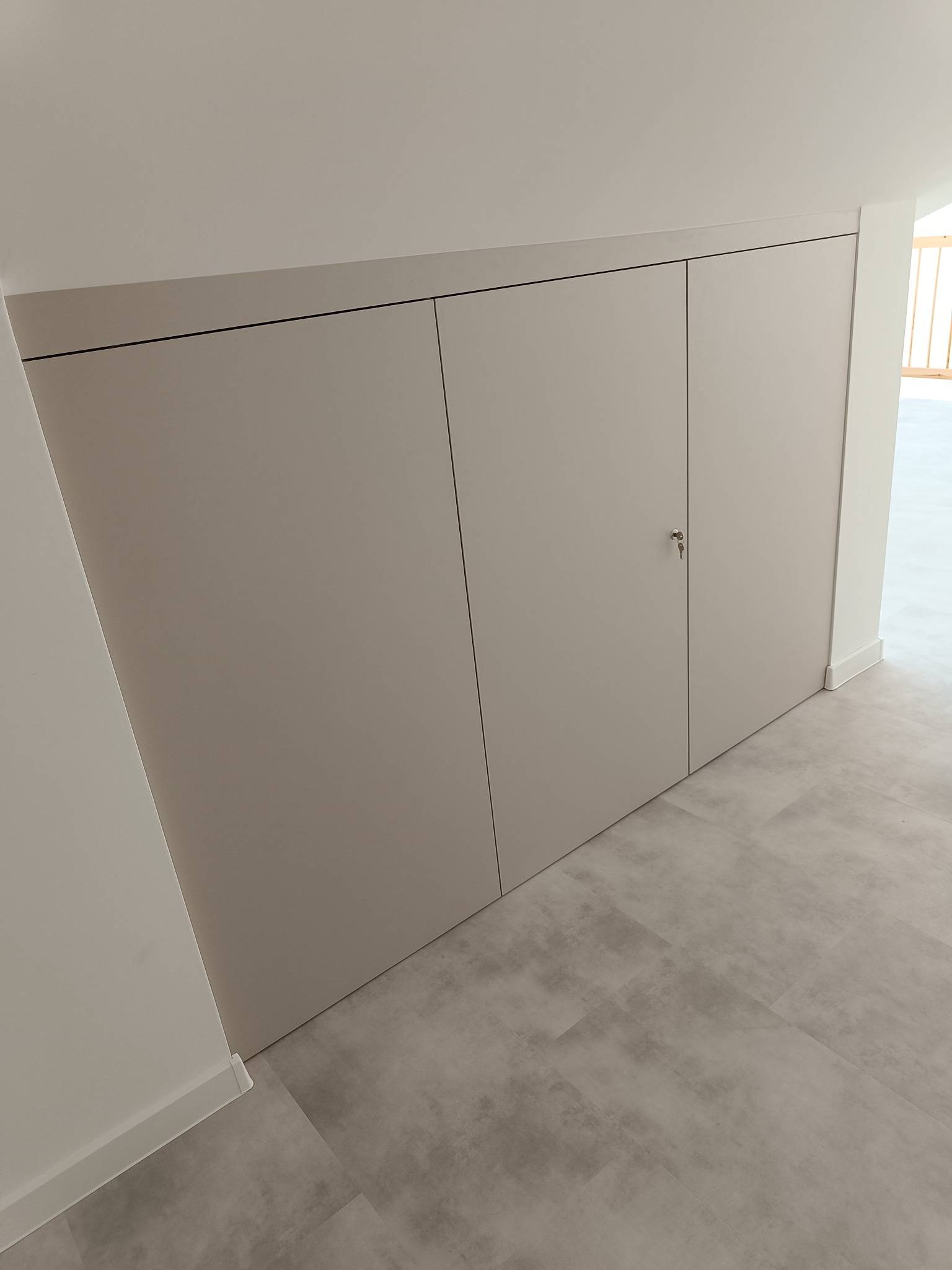 Jasna szafa wnękowa z trzema drzwiami, zamykana na klucz, wkomponowana w skos dachu, na tle szarej podłogi i białych ścian.