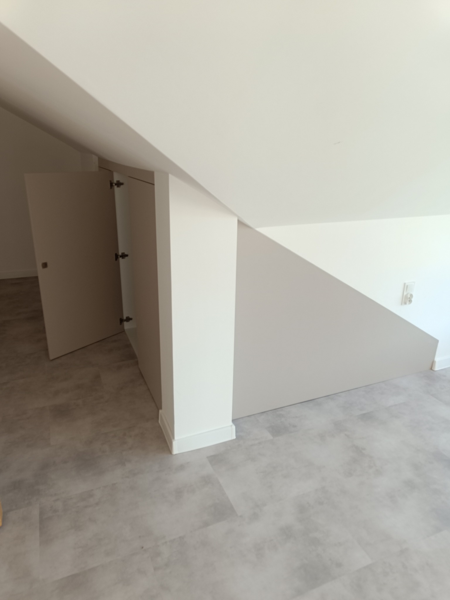 Biała szafa wnękowa z otwartymi drzwiami, dopasowana do skosu dachu, na szarej podłodze. Widoczne zawiasy i wnętrze szafy.