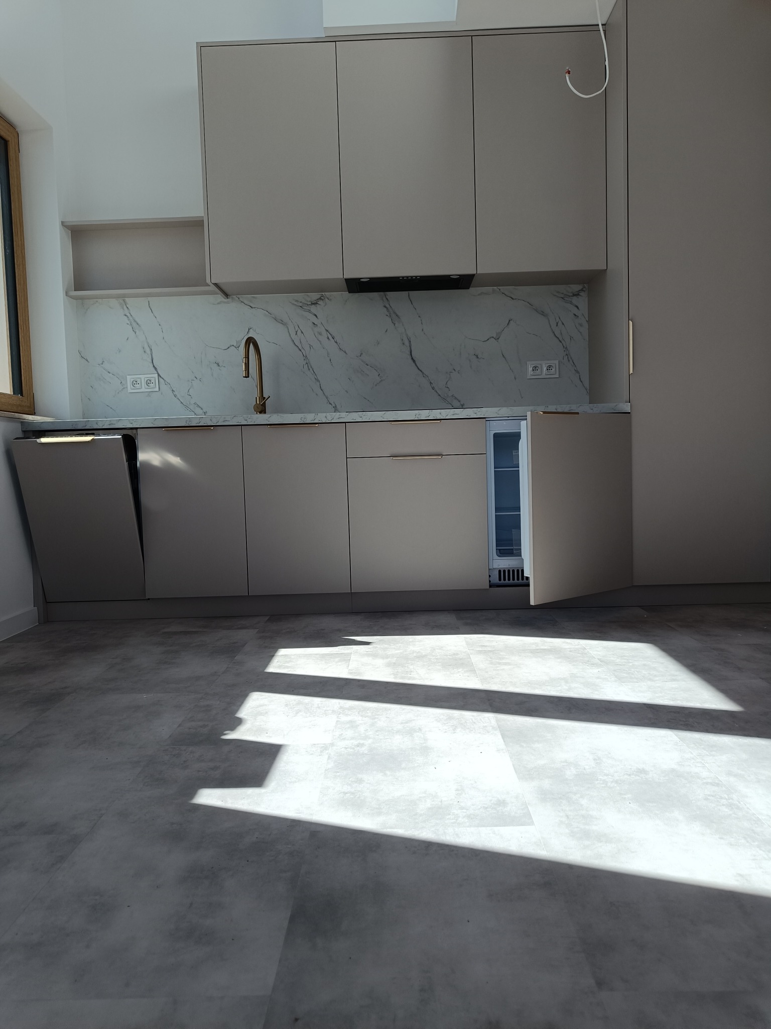 Jasna kuchnia z marmurowym blatem i złotą armaturą. Otwarta zmywarka i lodówka eksponują wnętrze. Na podłodze szare płytki, oświetlone promieniami słońca.