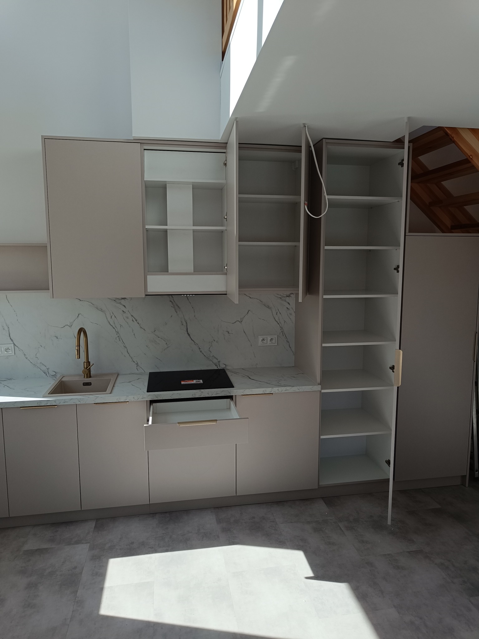 Jasna kuchnia na wymiar z marmurowym blatem i złotą armaturą. Częściowo otwarte szafki ukazują puste półki. Na podłodze widoczny cień.