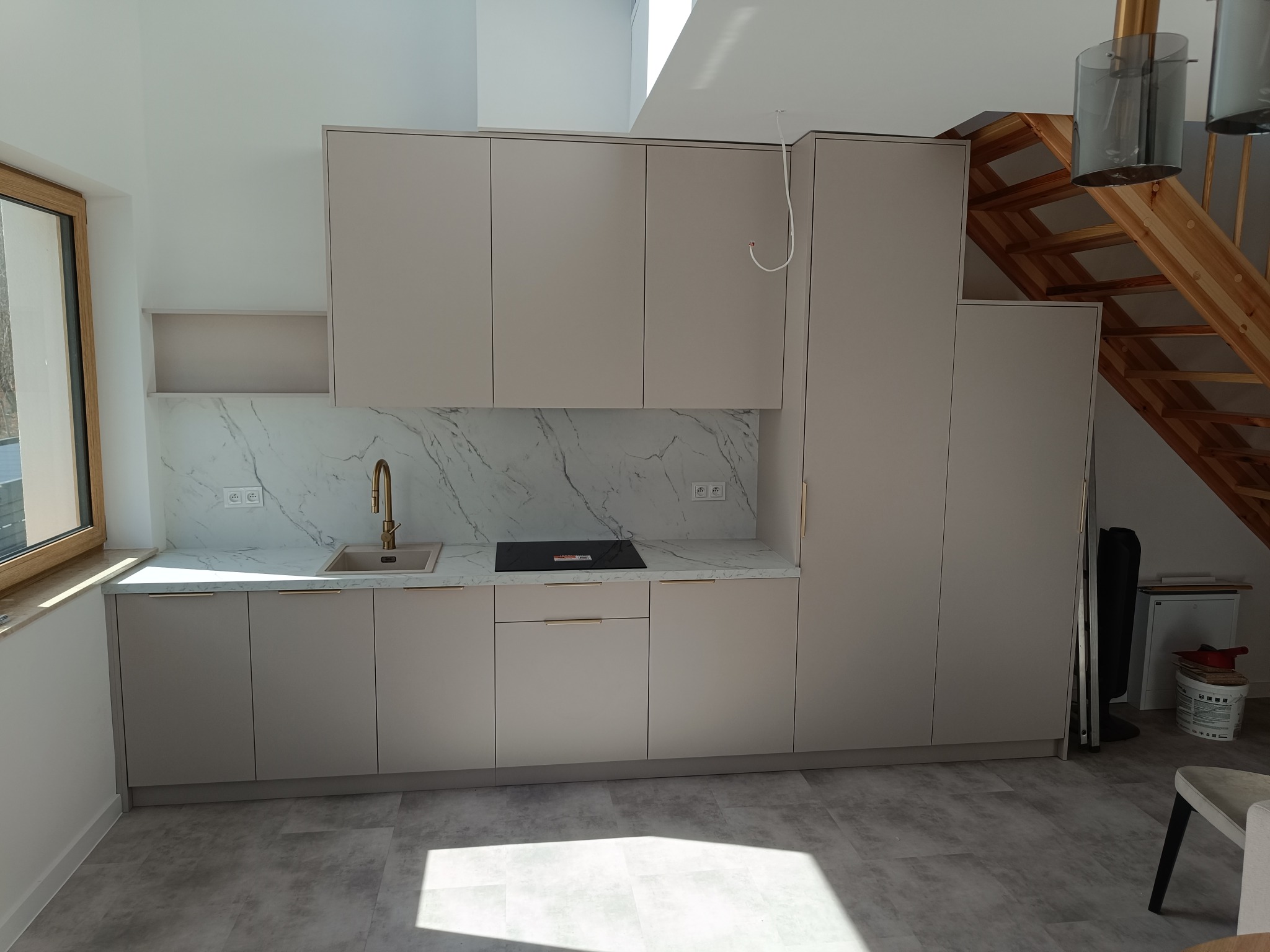 Jasna kuchnia na wymiar z marmurowym blatem i złotą armaturą, widok na drewniane schody i okno dachowe, minimalistyczny design.