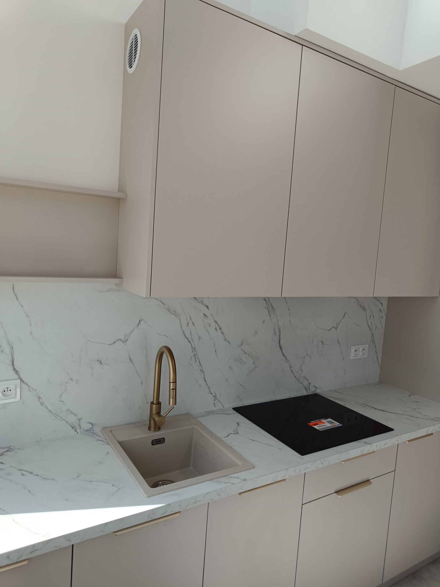 Jasna kuchnia na wymiar z marmurowym blatem, złotą baterią i płytą indukcyjną. Minimalistyczny design, gładkie fronty szafek i subtelne uchwyty.