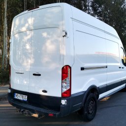 TRIOTRANS Paweł Czuba - Biały bus Ford Transit z widocznym hakiem holowniczym i tablicą rejestracyjną na tle drzew. Ujęcie z tyłu, lekko z boku, podkreślające przestrzeń ładunkową.
