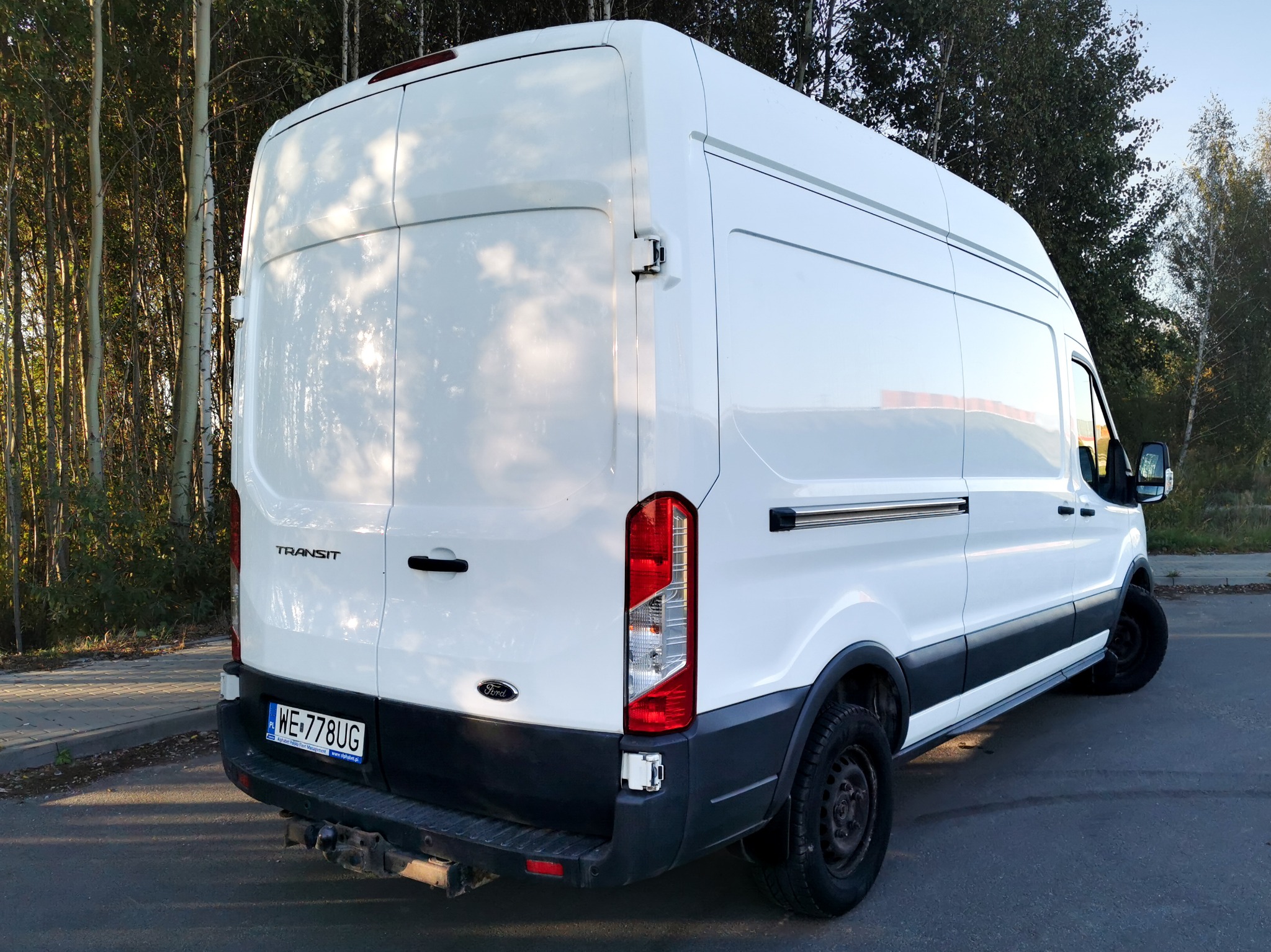Biały bus Ford Transit z widocznym hakiem holowniczym i tablicą rejestracyjną na tle drzew. Ujęcie z tyłu, lekko z boku, podkreślające przestrzeń ładunkową.