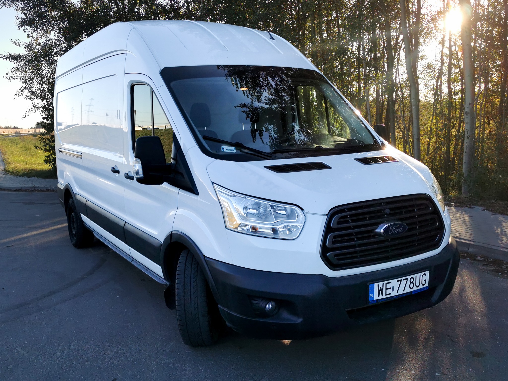 Biały Ford Transit z polską rejestracją WE-778UG, zaparkowany na asfalcie, z drzewami w tle i promieniami słońca. Widok z przodu, lekko z boku.