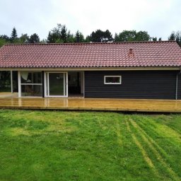 Łukasz Skorupski - Nowoczesny dom z ciemną elewacją i tarasem z jasnego drewna. Widoczne ślady kół na trawie. Dach z czerwonej dachówki. Osoba w tle przy samochodzie.