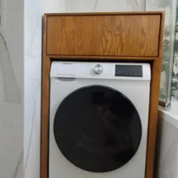 Biała pralka Hisense w drewnianej obudowie w łazience z marmurowymi płytkami. Obudowa z widocznym rysunkiem słojów drewna. Widok z przodu, lekko z dołu.