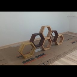 Łukasz Skorupski - Drewniane, heksagonalne półki w różnych odcieniach brązu i beżu, ułożone na podłodze zabezpieczonej kartonem, na tle białej ściany. Nietypowy kształt i kolorystyka.