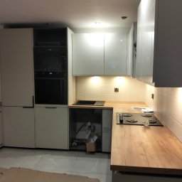 Łukasz Skorupski - Jasna kuchnia z drewnianym blatem i białymi szafkami. Widoczny zlew, płyta grzewcza i piekarnik. Na ścianie dekoracyjne, białe płytki. Pod szafką widoczne elementy instalacji.