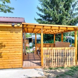 Łukasz Skorupski - Nowa drewniana konstrukcja: jasna szopa z metalowym dachem i dwoma lampami solarnymi, połączona z zadaszonym tarasem z kompozytową podłogą i ozdobną balustradą.