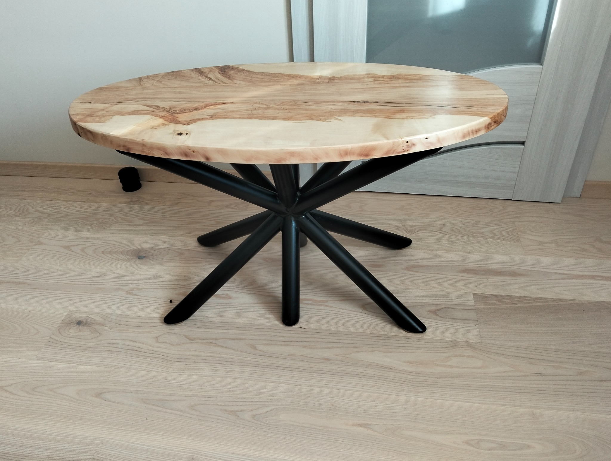 Okrągły stolik kawowy z blatem z jasnego drewna o unikalnym wzorze słojów i czarną, metalową podstawą o geometrycznym kształcie, na tle podłogi z paneli.