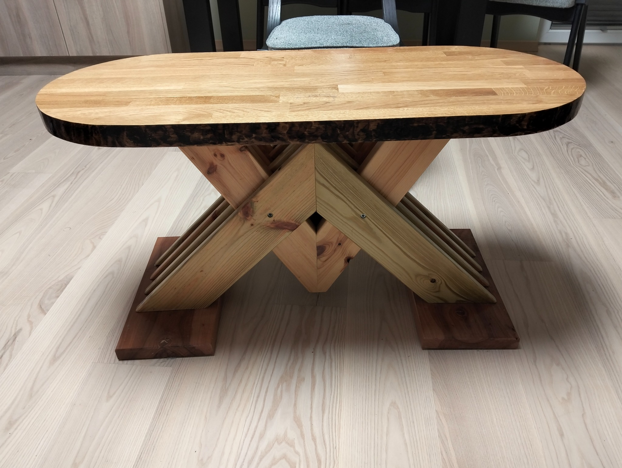 Owalny stolik kawowy z blatem z jasnego drewna o nieregularnej krawędzi, wsparty na konstrukcji z krzyżujących się belek i ciemnych podstawach, na jasnej podłodze.