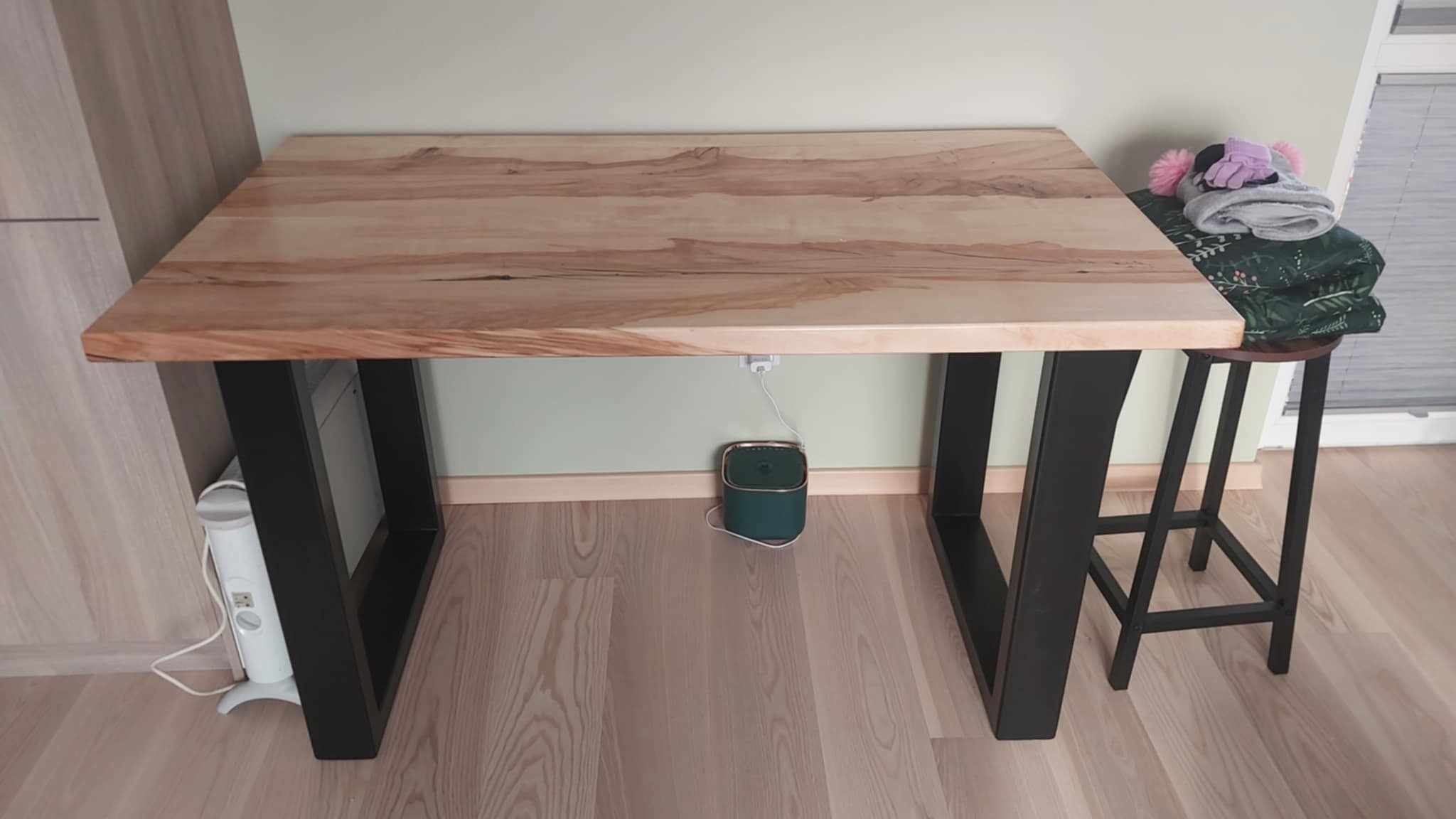 Stół z blatem z jasnego drewna o wyraźnym usłojeniu, wsparty na czarnych, metalowych nogach. W tle ściana w odcieniu zieleni i fragment grzejnika.