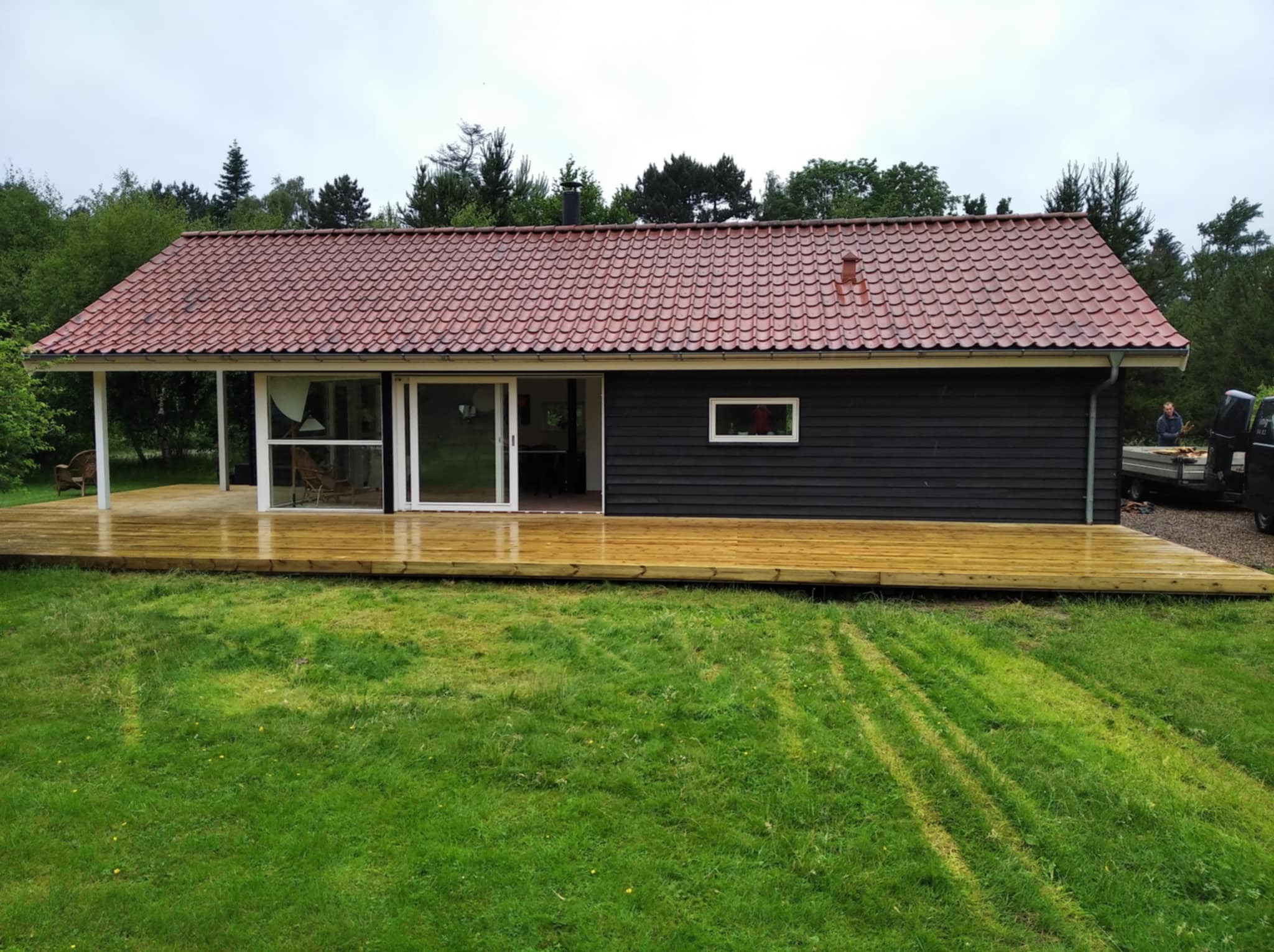 Nowoczesny dom z ciemną elewacją i tarasem z jasnego drewna. Widoczne ślady kół na trawie. Dach z czerwonej dachówki. Osoba w tle przy samochodzie.