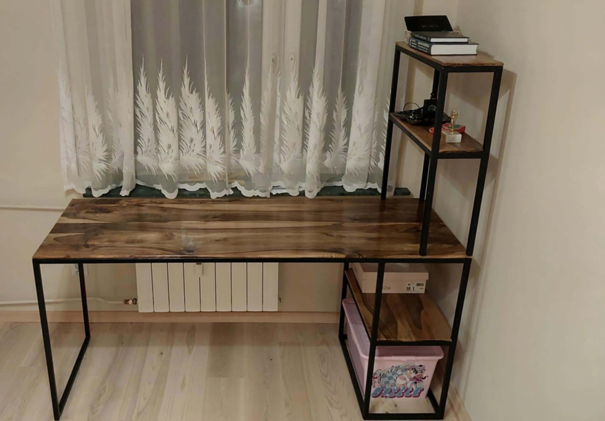 Drewniane biurko z metalowym stelażem, z półkami po prawej stronie, na których leżą książki i pudełka. W tle okno z firanką w motywy roślinne.