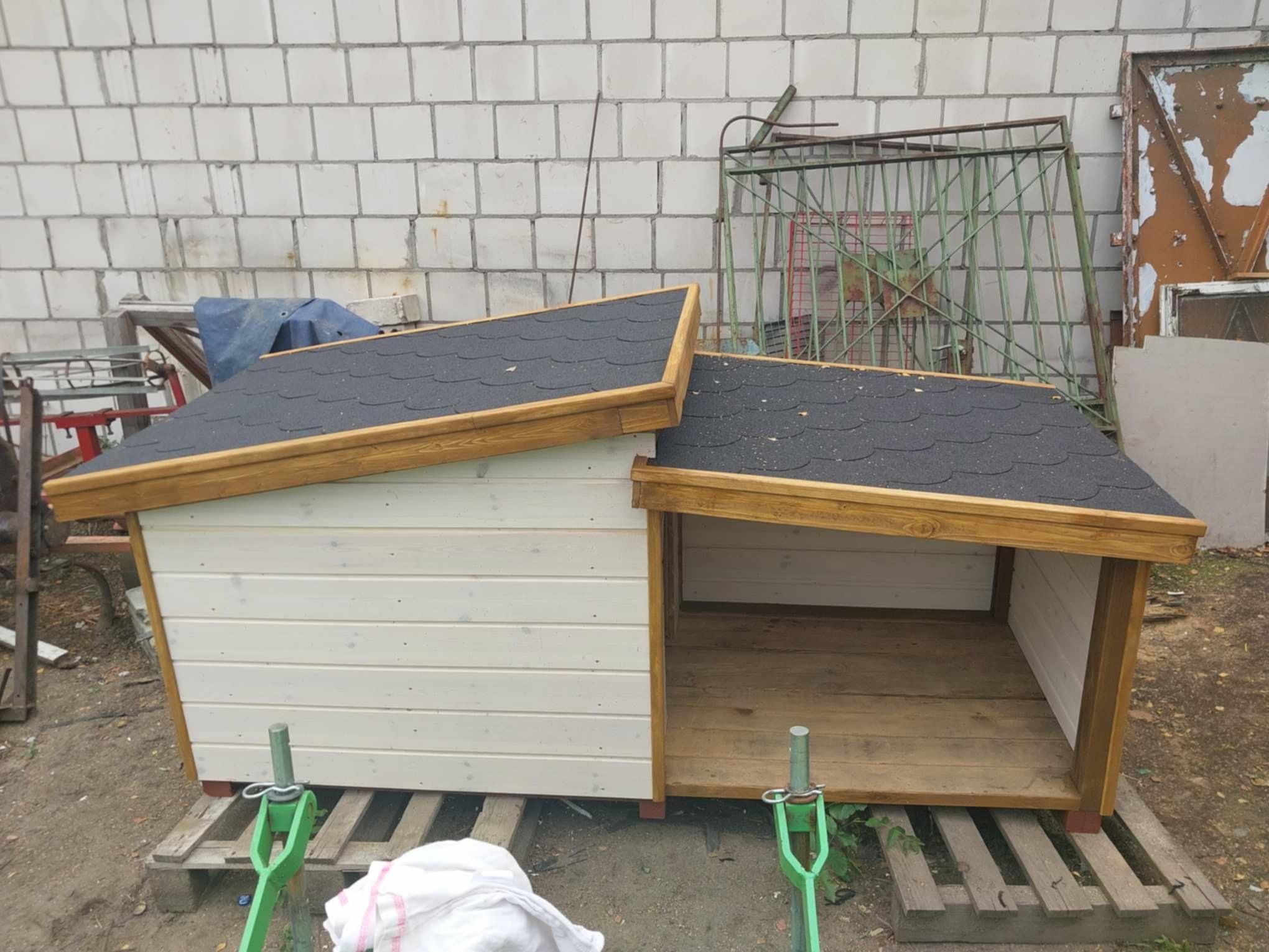 Drewniana buda dla psa z dwuspadowym dachem pokrytym papą, pomalowana na biało i brązowo, stoi na palecie przed ścianą z bloczków.