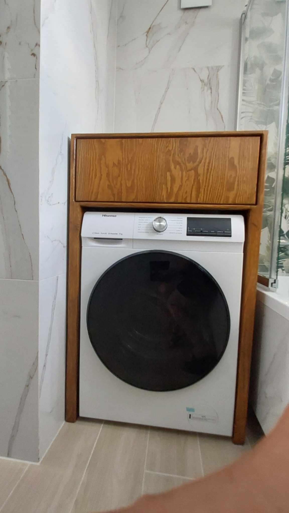 Biała pralka Hisense w drewnianej obudowie w łazience z marmurowymi płytkami. Obudowa z widocznym rysunkiem słojów drewna. Widok z przodu, lekko z dołu.