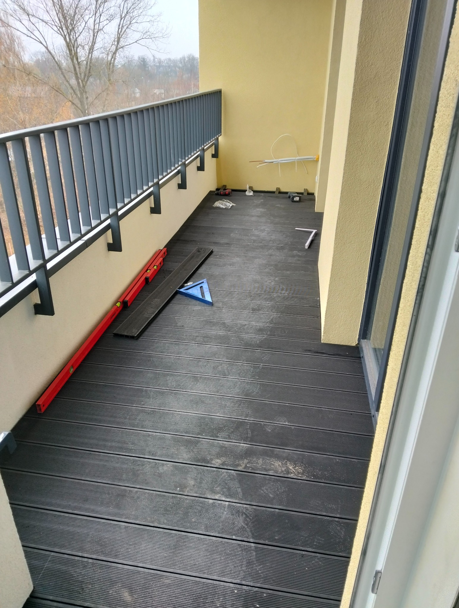 Montaż deski kompozytowej na balkonie. Widoczne poziomice, kątownik i narzędzia. Częściowo ułożona podłoga z desek w kolorze grafitowym. Żółta ściana i szara balustrada.