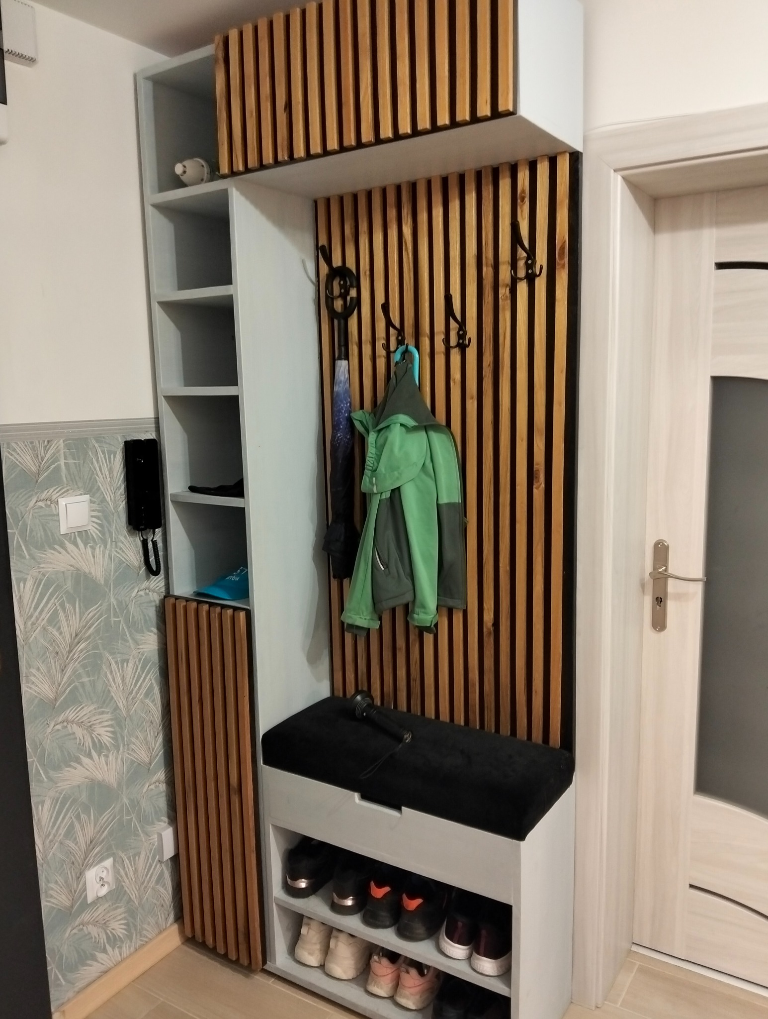 Funkcjonalna zabudowa przedpokoju z drewnianymi lamelami, szafką na buty z siedziskiem i wieszakami na ubrania. Nowoczesny design, szara kolorystyka.