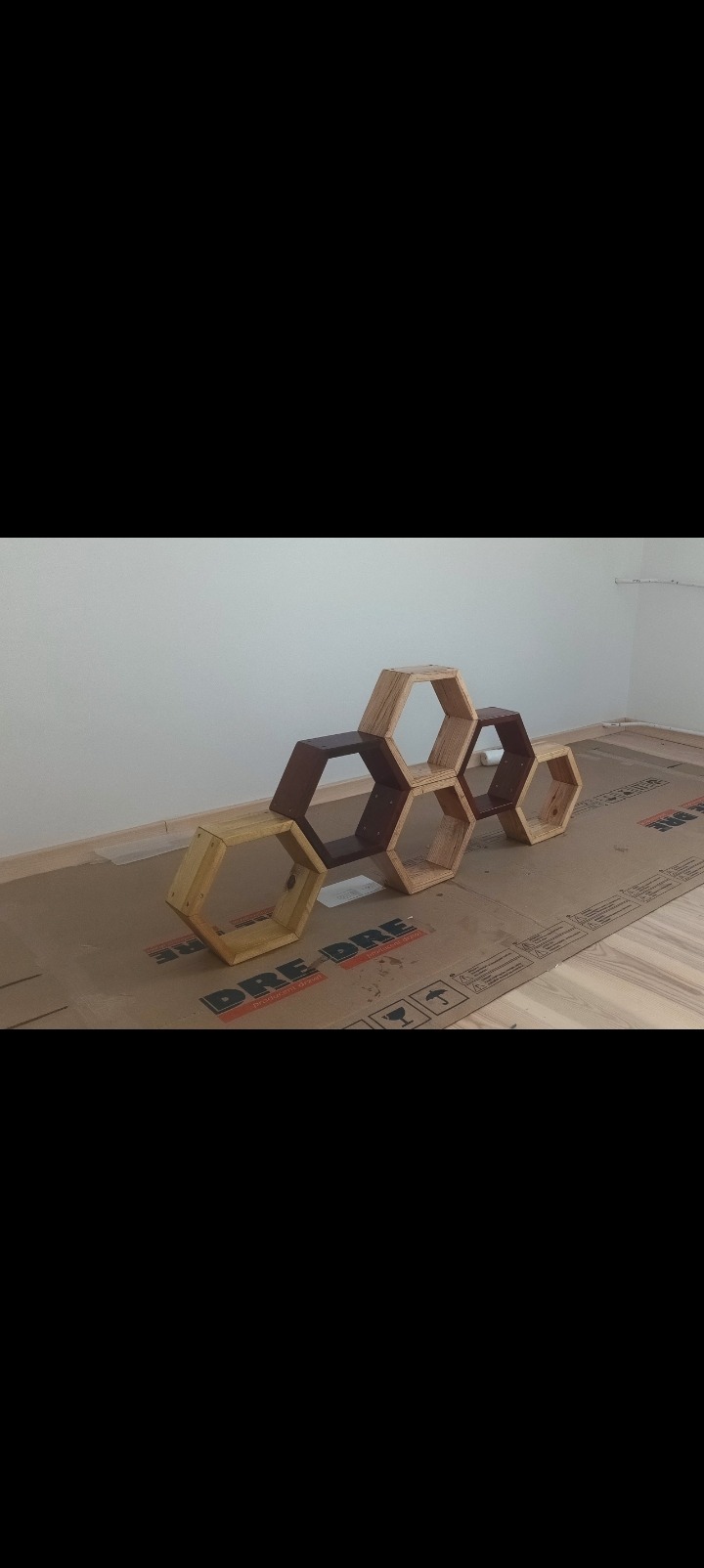 Drewniane, heksagonalne półki w różnych odcieniach brązu i beżu, ułożone na podłodze zabezpieczonej kartonem, na tle białej ściany. Nietypowy kształt i kolorystyka.