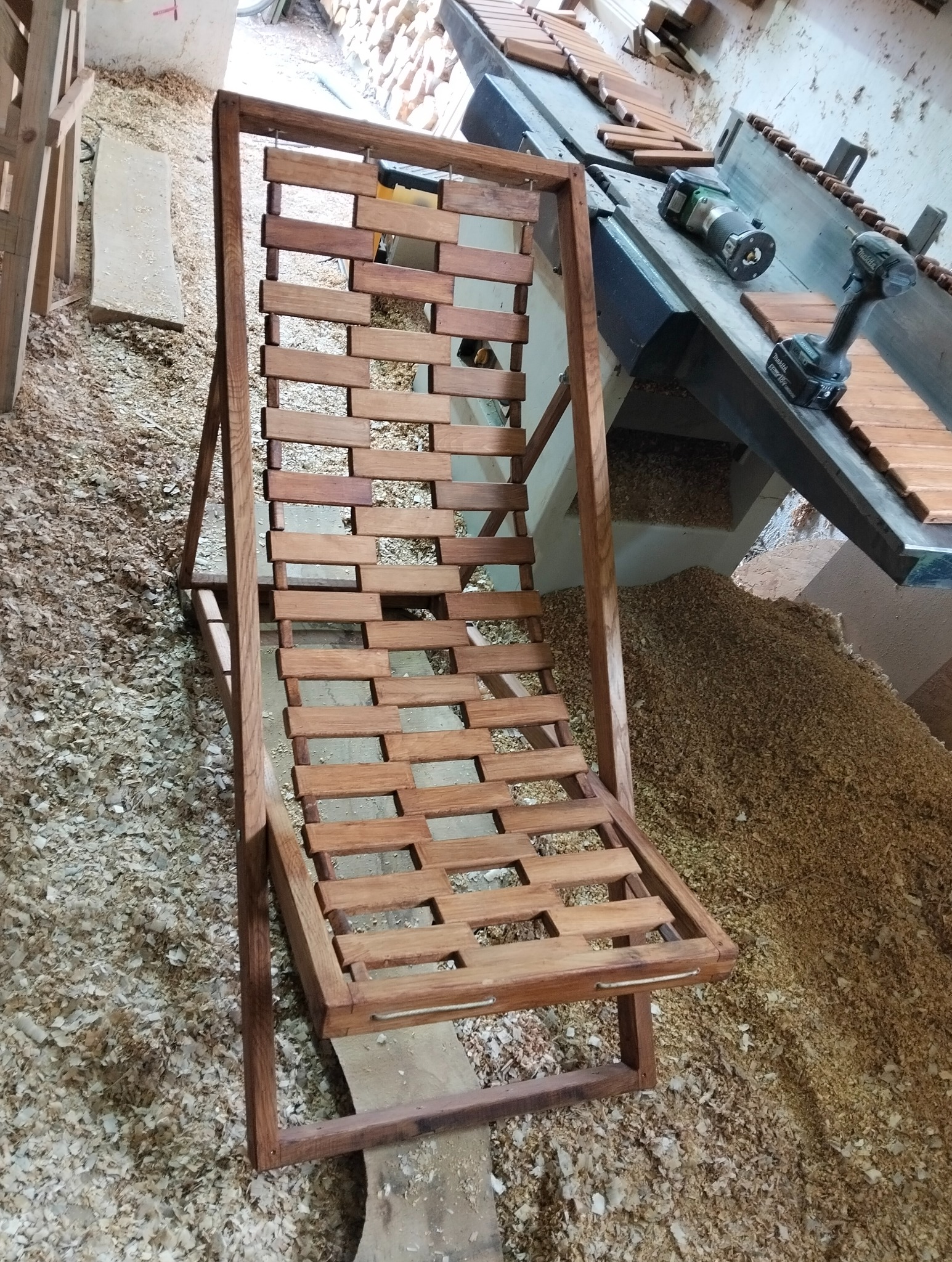 Drewniany, ażurowy leżak w trakcie produkcji, widoczne narzędzia stolarskie i sterta wiórów. Ujęcie z góry, ukazujące detale konstrukcji i warsztatowe otoczenie.