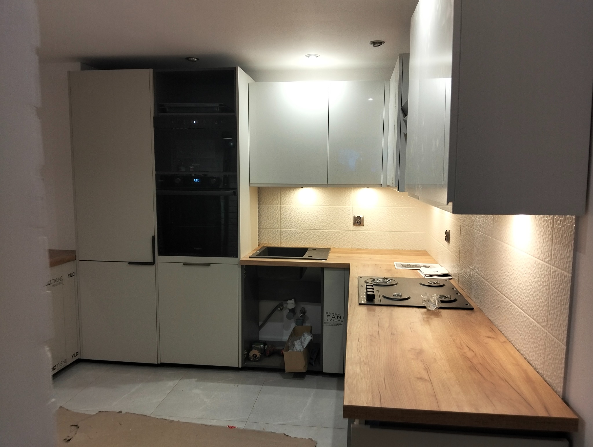 Jasna kuchnia z drewnianym blatem i białymi szafkami. Widoczny zlew, płyta grzewcza i piekarnik. Na ścianie dekoracyjne, białe płytki. Pod szafką widoczne elementy instalacji.