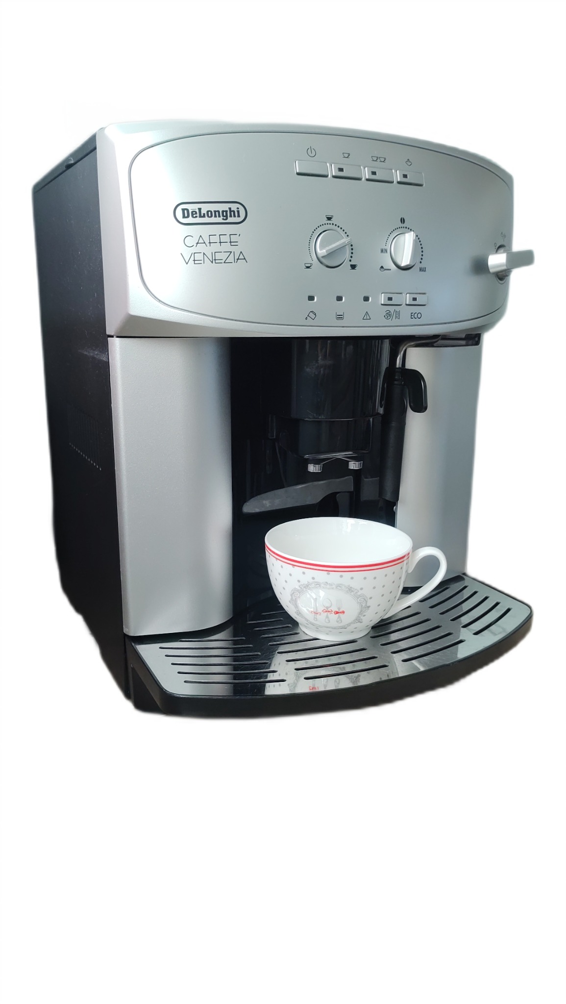 Szaro-czarny ekspres DeLonghi Caffe Venezia z filiżanką na ociekaczu. Widoczne pokrętła regulacji i ikony funkcji. Górna część obudowy srebrna, bok czarny.