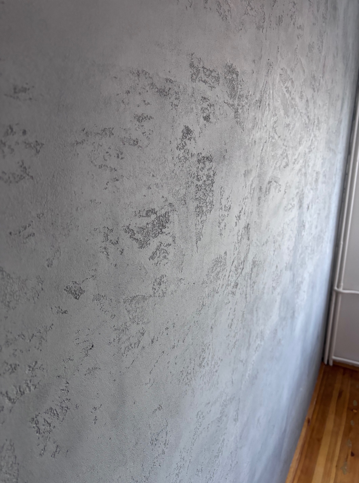 Ściana z dekoracyjną, szarą fakturą, imitującą efekt betonu architektonicznego. Widoczny fragment drewnianej podłogi i biała listwa przypodłogowa.