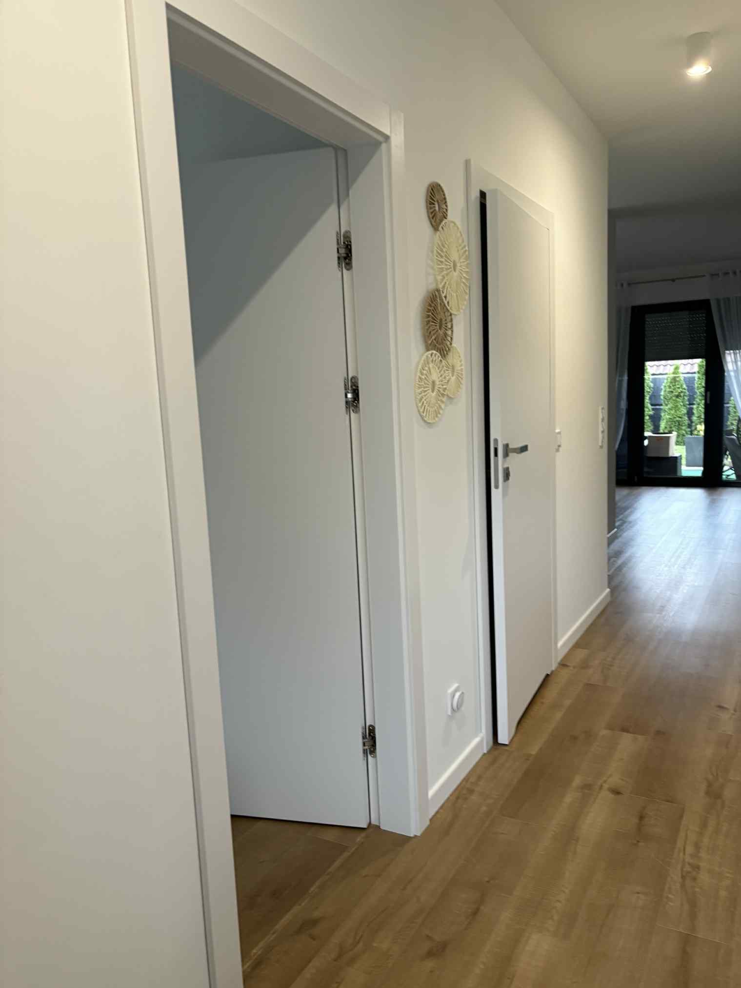 Jasny korytarz z drewnianą podłogą i białymi drzwiami. Na ścianie dekoracyjne, okrągłe ozdoby. Widok na ogród przez otwarte drzwi balkonowe.