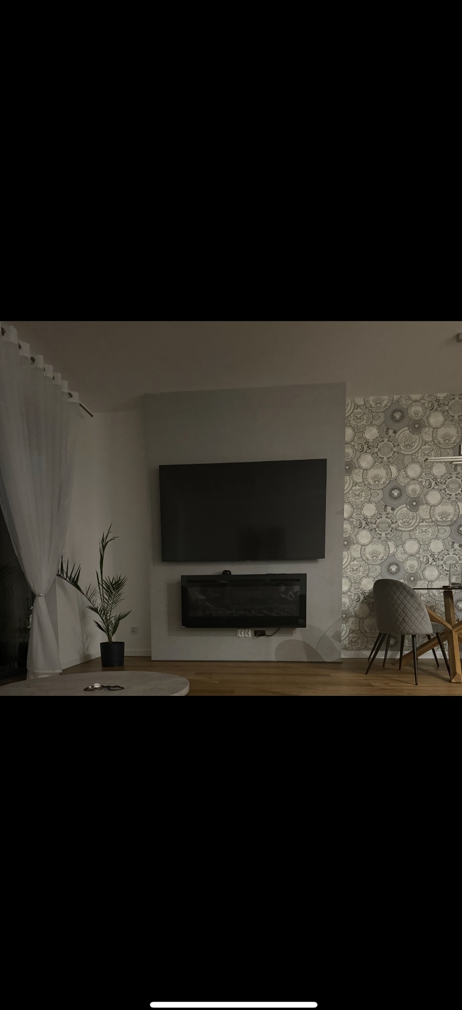 Salon z telewizorem i kominkiem elektrycznym pod nim. Na ścianie wzorzysta tapeta. Minimalistyczne wnętrze z rośliną w doniczce i okrągłym stolikiem kawowym.